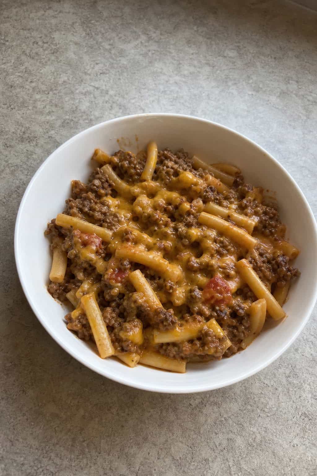 A bowl of cheesy beef and pasta dinner garnished with parsley on a wooden table with a fork and napkin nearby.