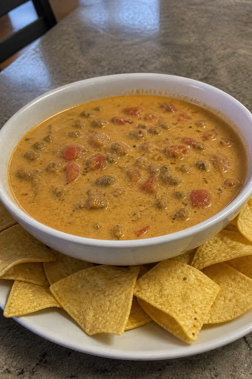 A bowl of cheesy chili con queso with ground beef and diced tomatoes surrounded by tortilla chips on a wooden table.