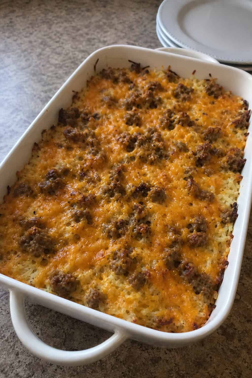 A freshly baked hashbrown casserole with crumbled sausage and melted cheese in a white ceramic dish, sitting on a wood table next to a glass of orange juice and a bowl of fresh berries.