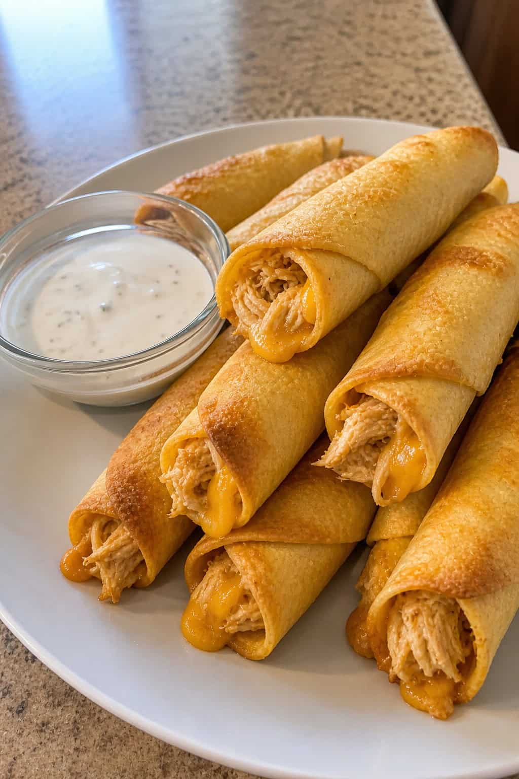 A plate of crispy cheesy ranch chicken taquitos with a bowl of ranch dipping sauce on a wooden table.