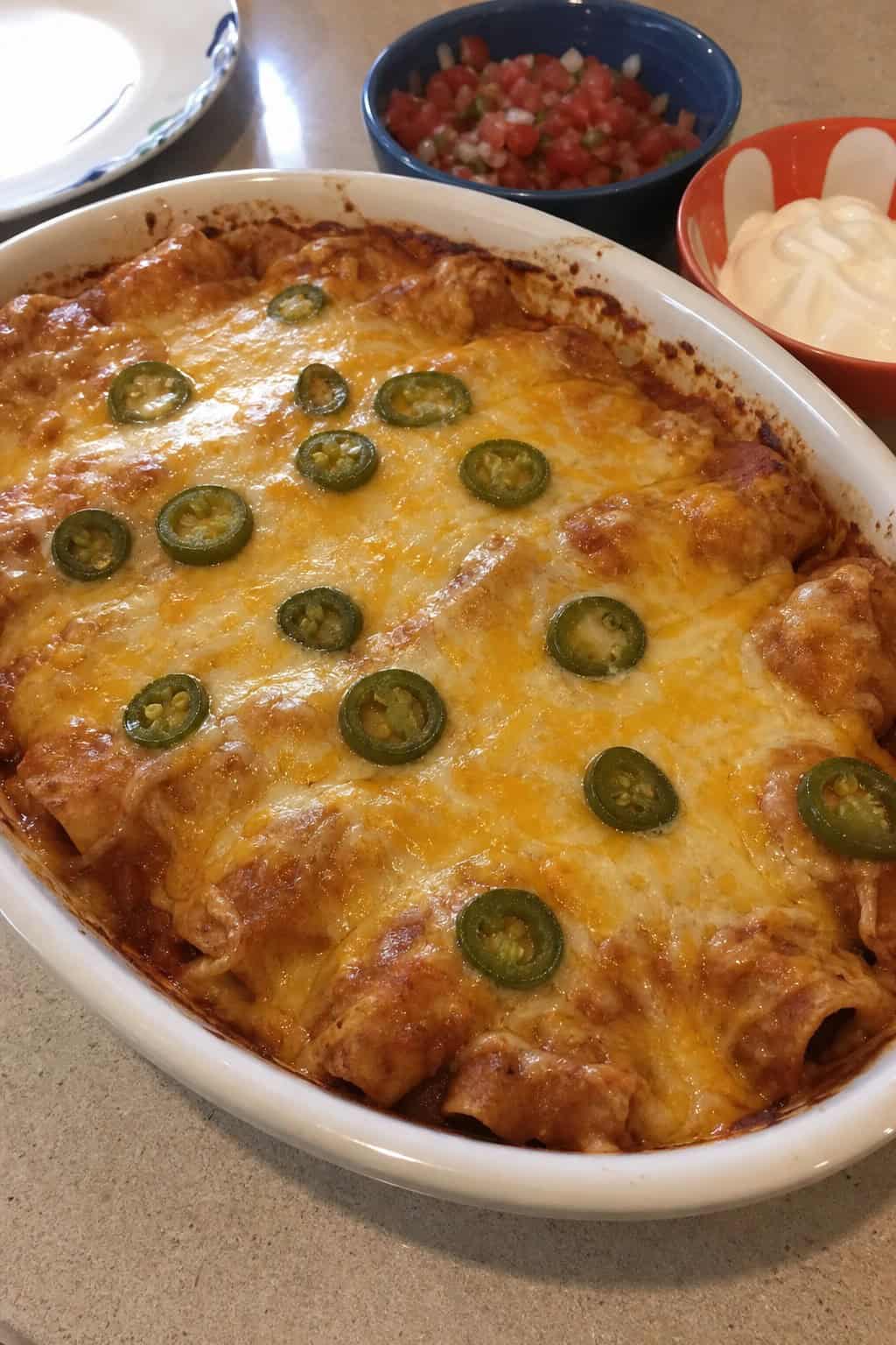 A baked rotisserie chicken enchilada casserole in a white ceramic dish with deep golden melted cheese on top, garnished with fresh cilantro and sliced jalapeños, with small bowls of salsa and sour cream nearby.