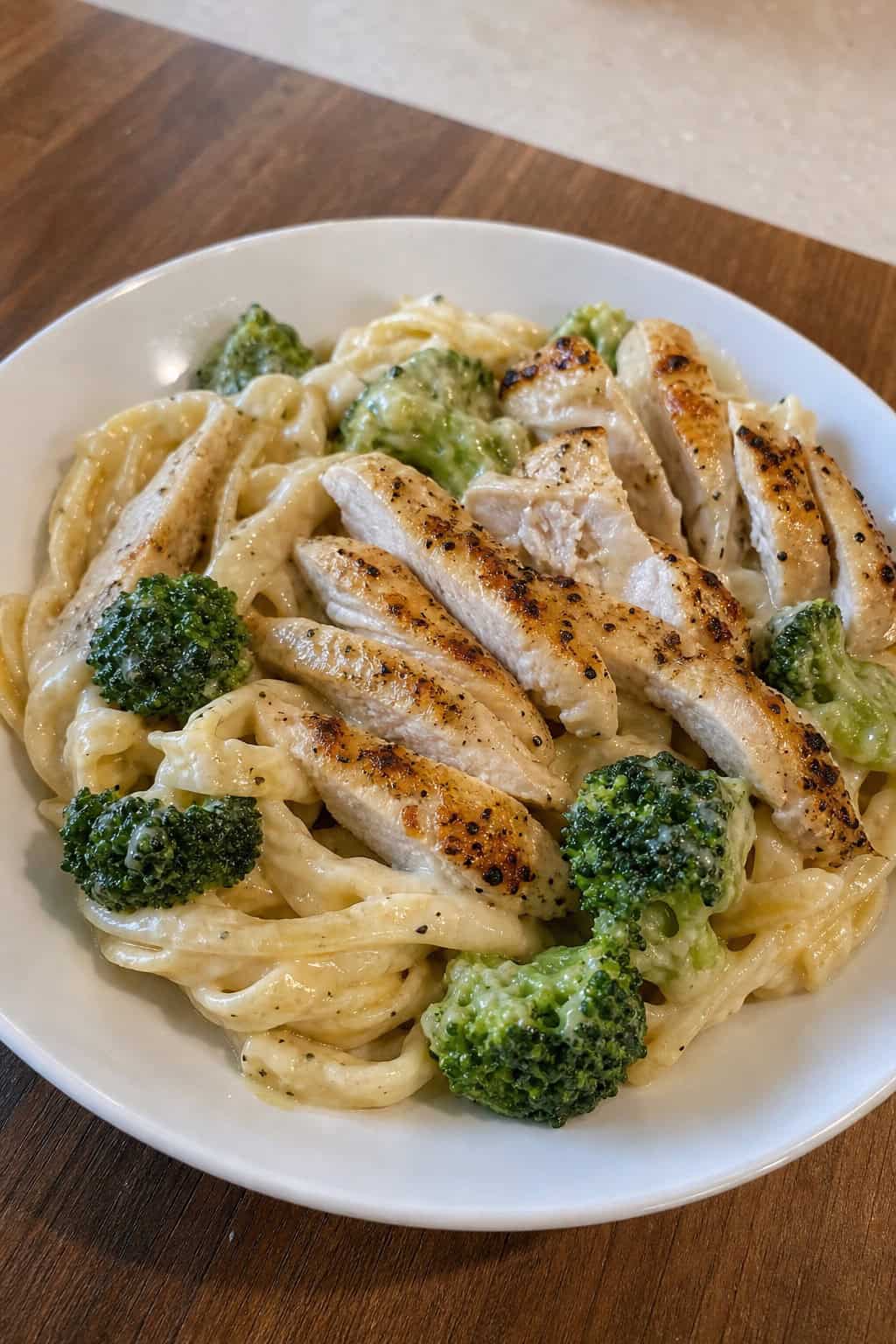A plate of chicken Alfredo pasta topped with broccoli on a wooden table.