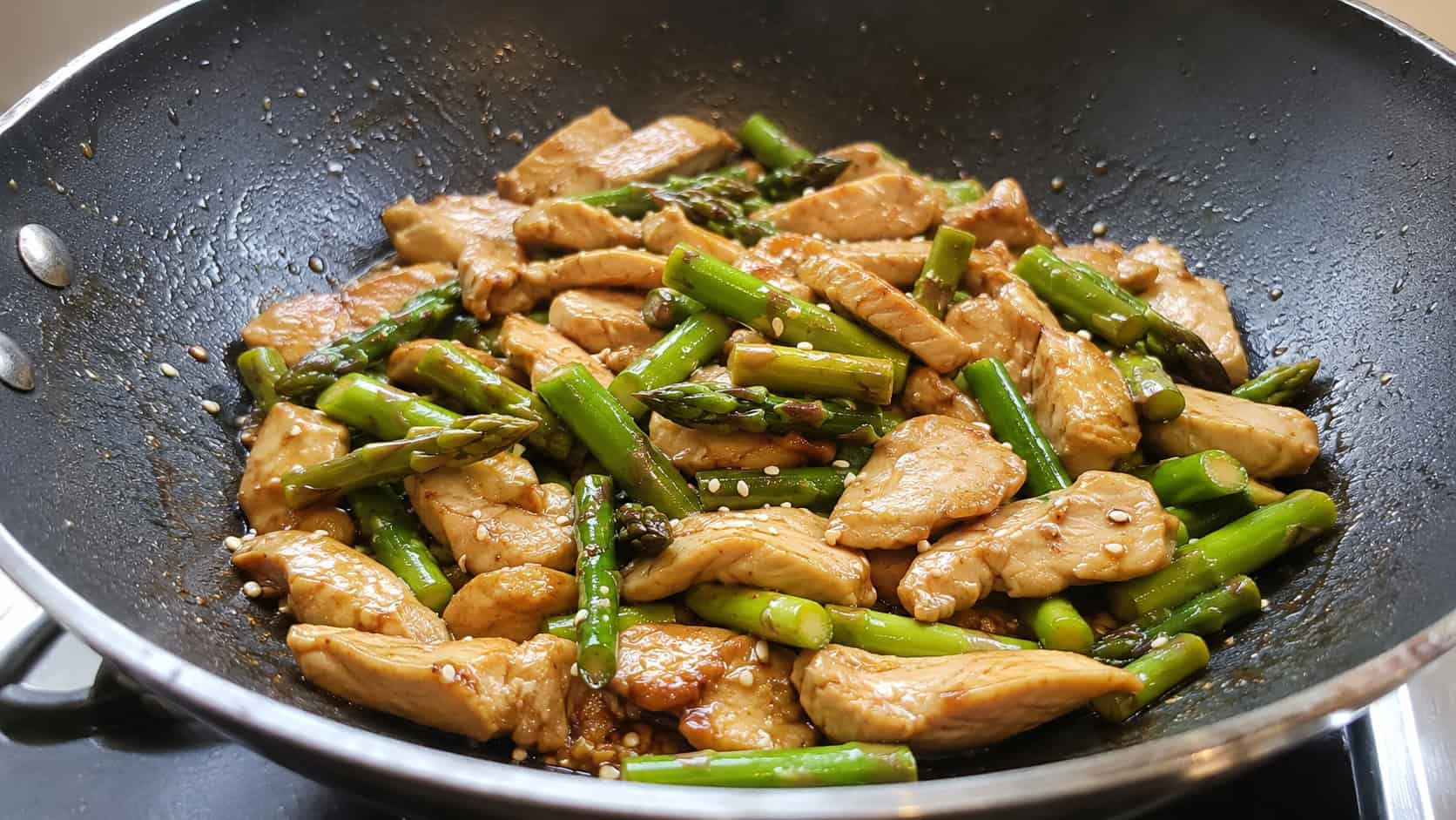 25 Heart-Healthy Chicken Recipes to Keep You Feeling Great! 26 a finished chicken and asparagus stir fry in a wok, sliced chicken and bright green asparagus pieces glazed in a light glossy sauce, sesame seeds scattered on top, warm natural kitchen light, casual home cooking