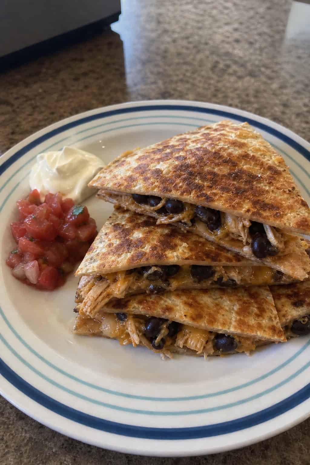 A wooden cutting board with golden-brown quesadilla wedges, garnished with fresh cilantro, sliced jalapeños, lime wedges, and small bowls of salsa and sour cream alongside.