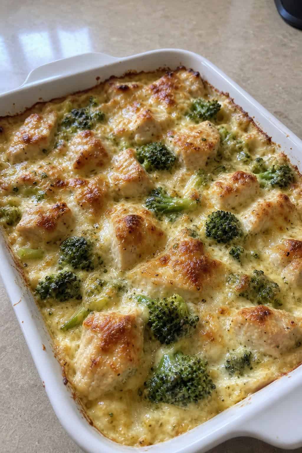 A baked chicken and broccoli Alfredo casserole in a white dish on a wooden table.