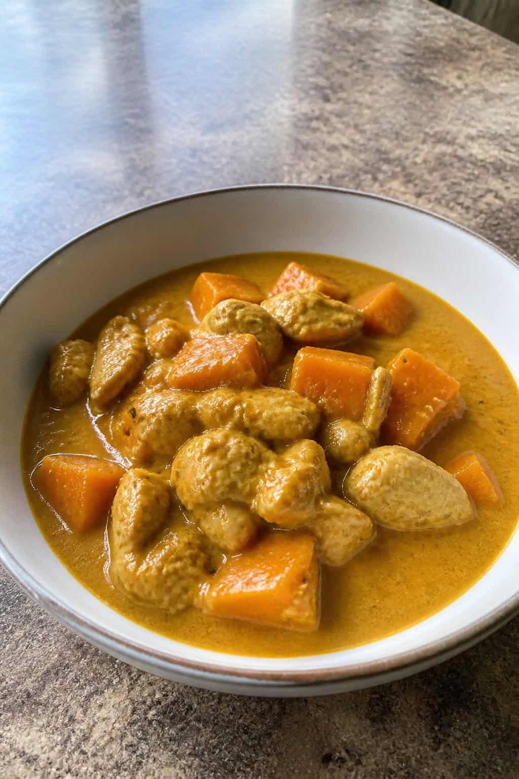 A bowl of chicken and butternut squash curry garnished with cilantro on a wooden table with fresh ingredients around it.