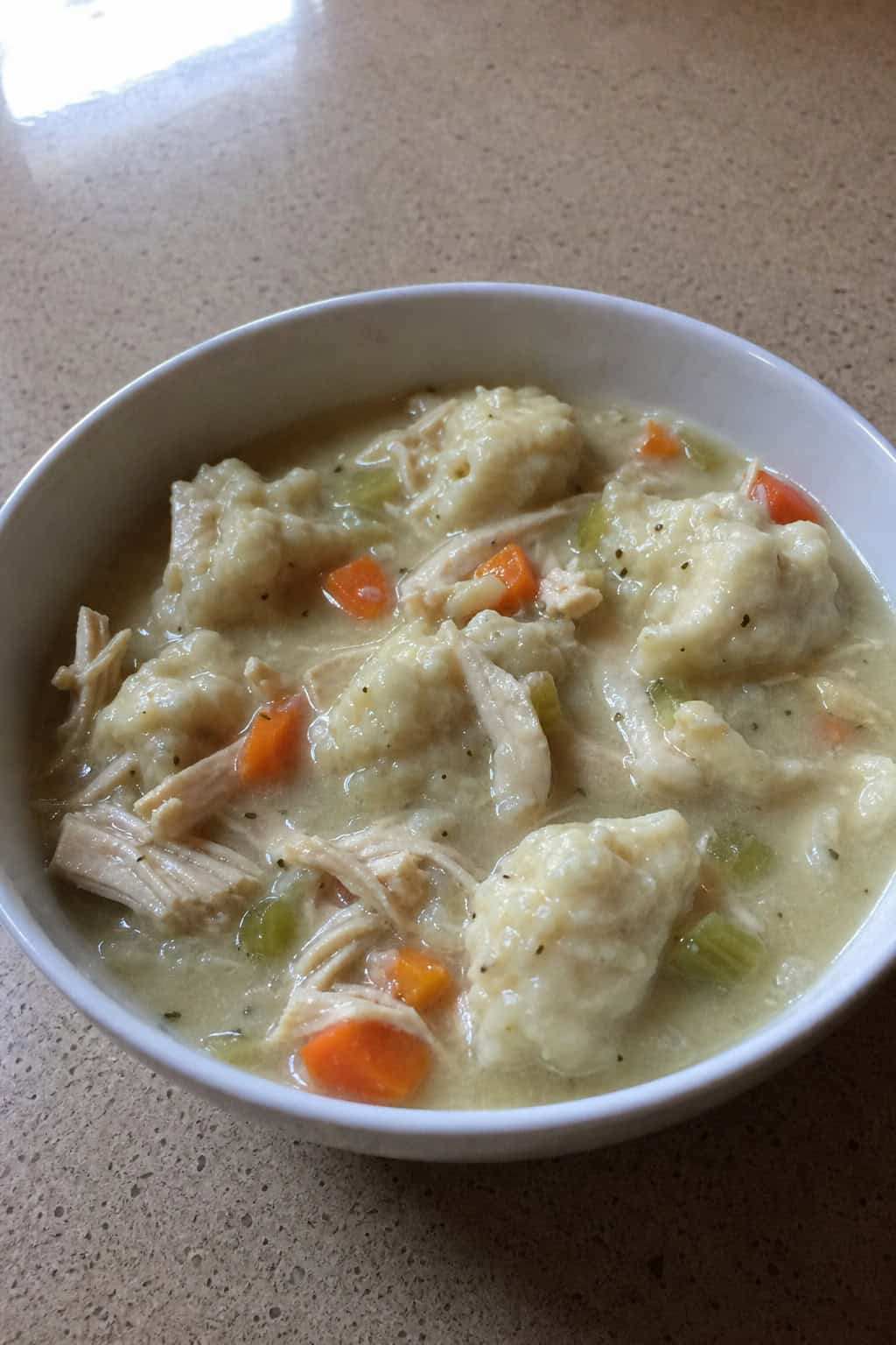 A steaming bowl of chicken and dumplings on a wooden table with fresh herbs and ingredients nearby.