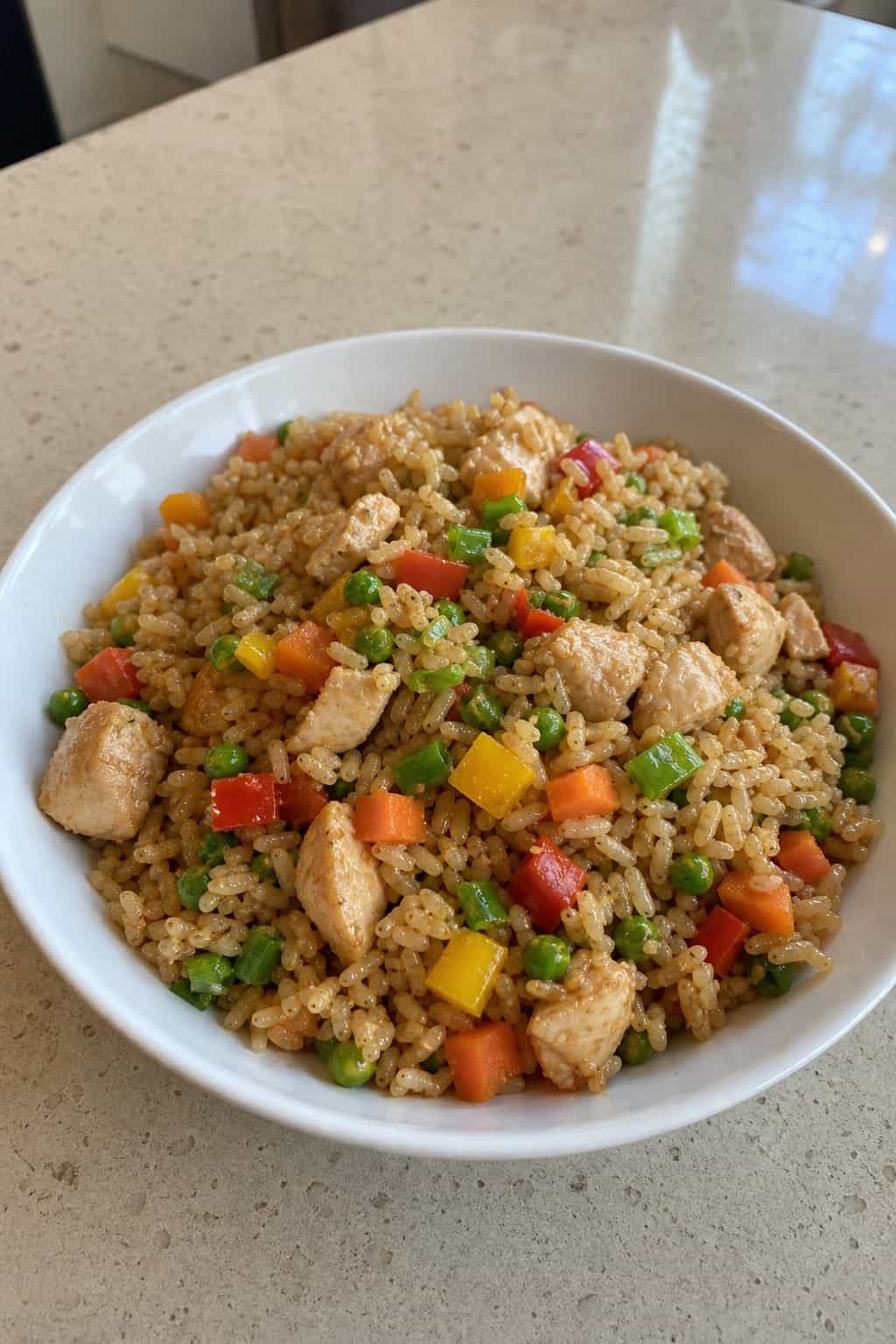 A bowl of chicken and vegetable fried rice with brown rice on a wooden table, surrounded by fresh vegetables.