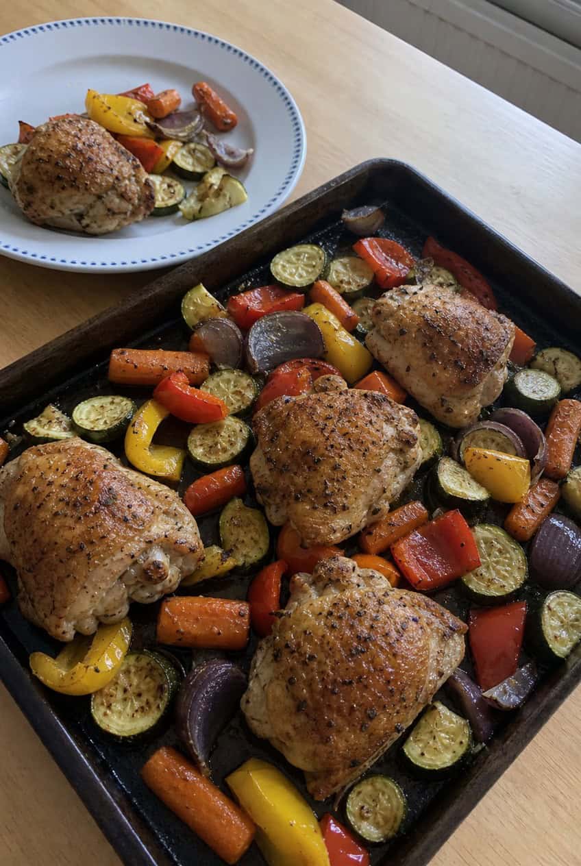 A foil-lined sheet pan with four seasoned chicken thighs surrounded by roasted broccoli, halved baby potatoes, and red bell pepper slices on a wooden surface.
