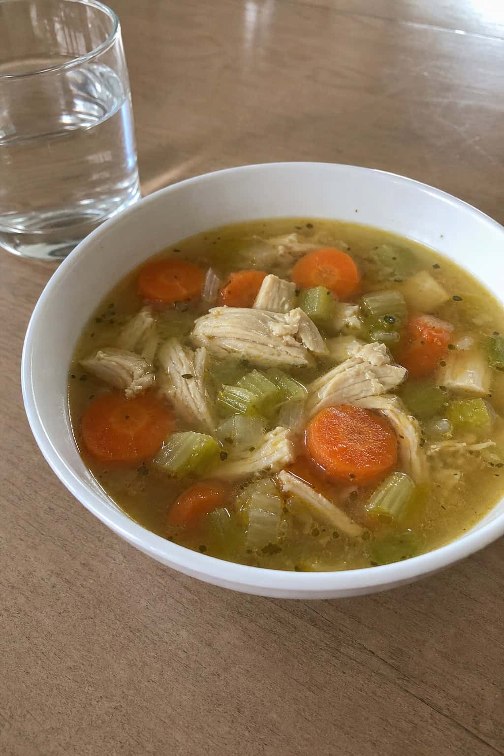 A steaming bowl of chicken and vegetable soup with fresh ingredients on a wooden table.