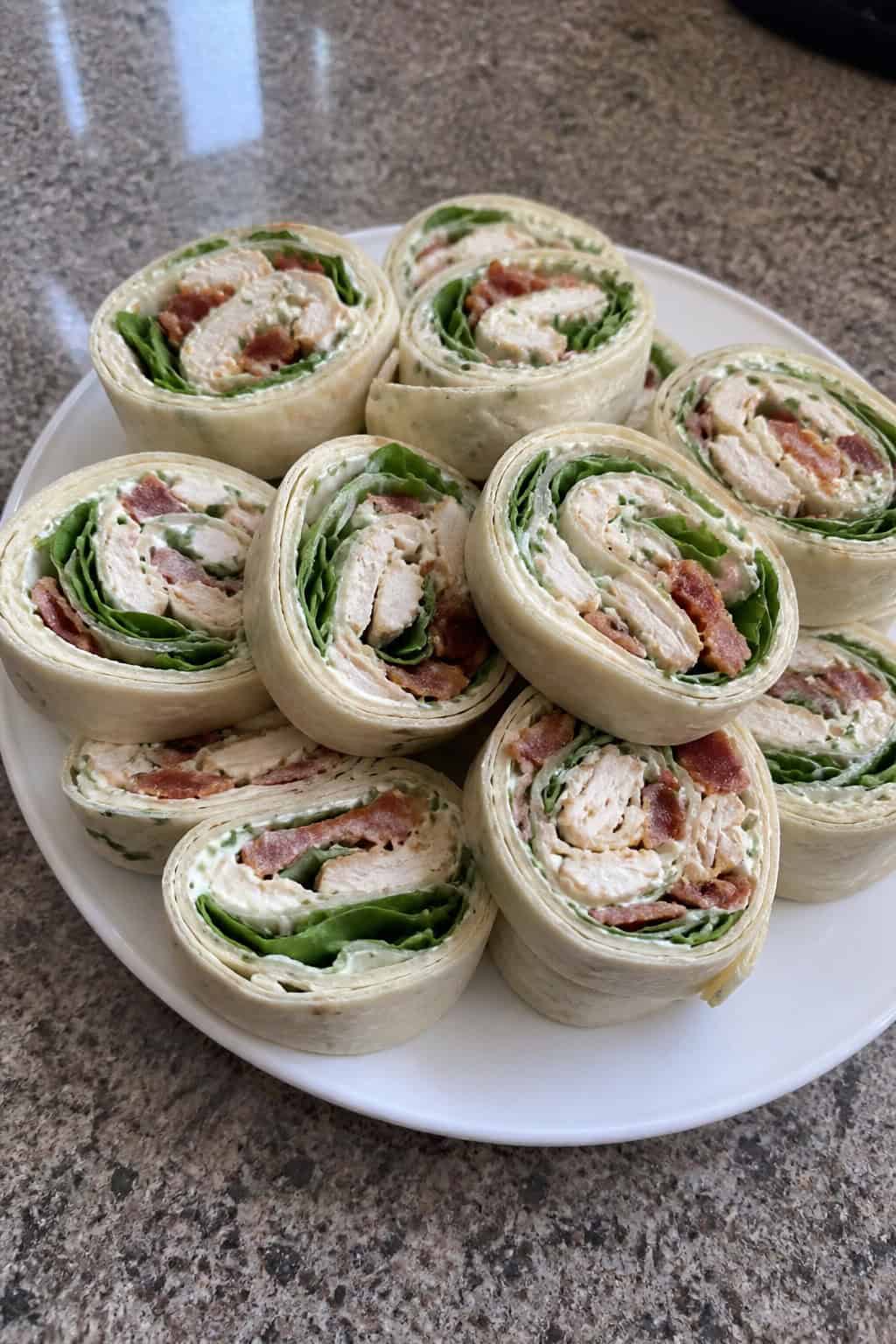 Close-up of sliced chicken bacon ranch pinwheels arranged on a wooden board showing shredded chicken, chopped bacon, and cheddar in a creamy ranch filling, with a small dipping sauce bowl in the background.