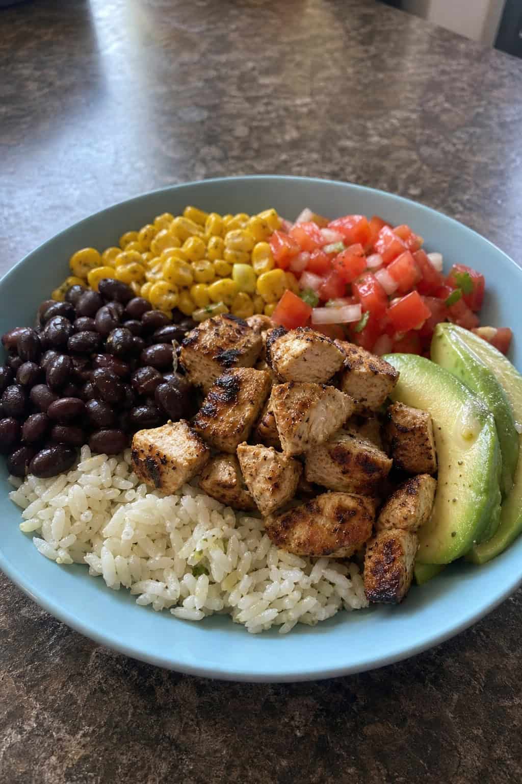 A wide ceramic bowl holding cilantro lime rice topped with shredded spiced chicken, sliced avocado, black beans, yellow corn, diced tomatoes, and a lime wedge on a wooden table.