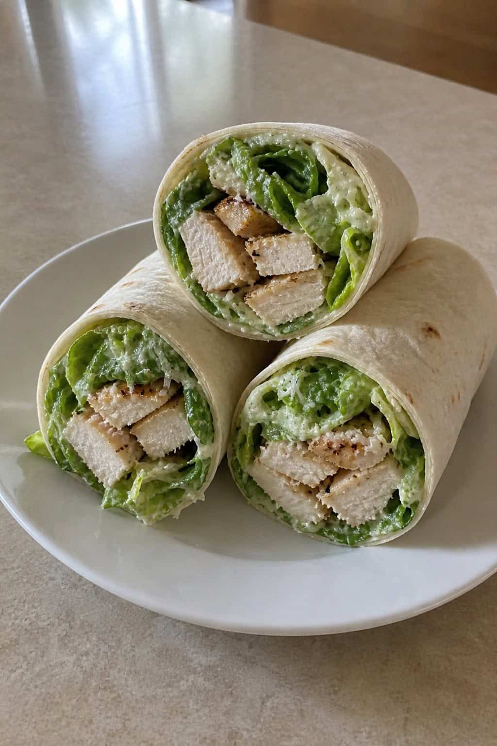 Close-up of two halved chicken Caesar wraps on a wooden cutting board, showing the filling of shredded chicken, thinly sliced romaine, and crushed croutons wrapped in a flour tortilla.