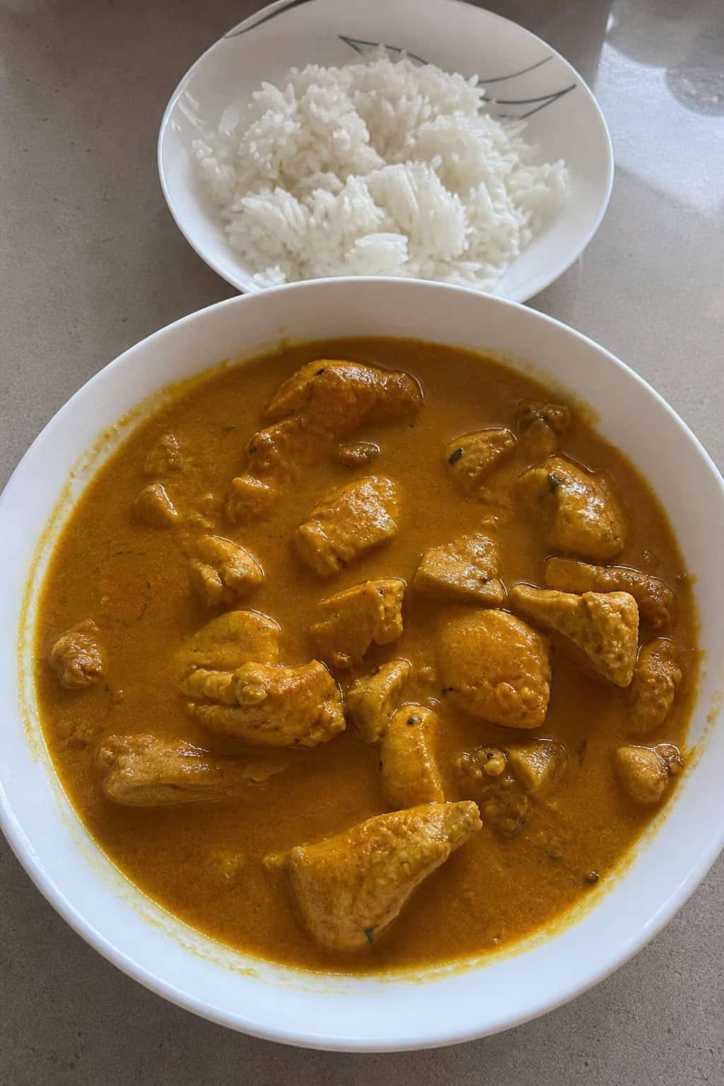 A bowl of creamy chicken curry with coconut milk garnished with cilantro on a wooden table with fresh ingredients around it.