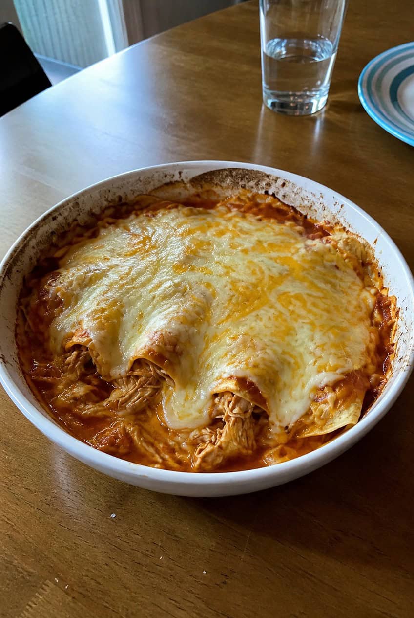 A just-baked chicken enchilada casserole with bubbling melted cheese and scattered fresh cilantro on top, set on a wooden table next to a small bowl of red salsa and a halved lime.