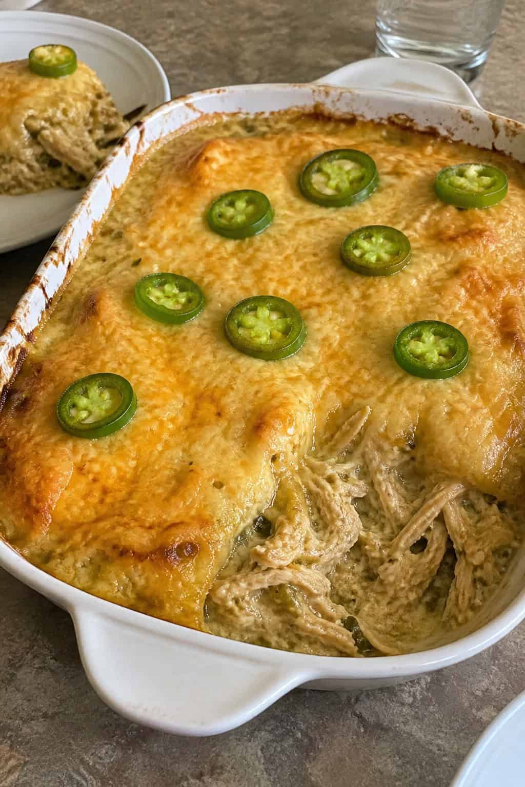 A baked chicken enchilada casserole with melted Monterey Jack cheese and diced green chili in a ceramic dish, garnished with fresh cilantro and sliced jalapeños on a wooden table.