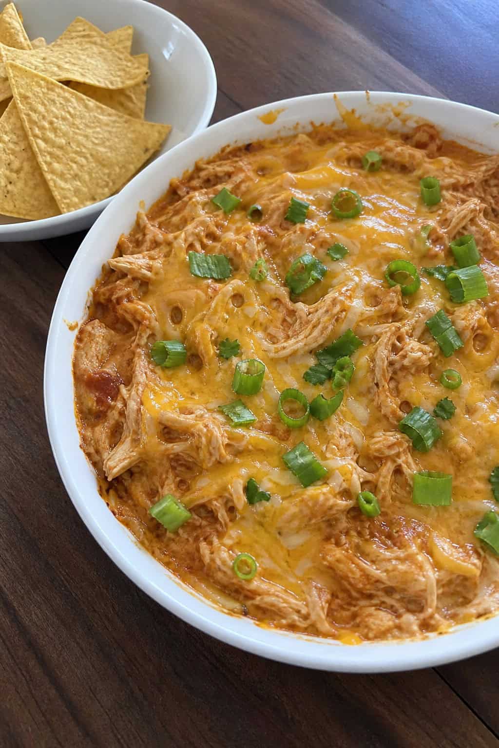 A bowl of chicken enchilada dip with shredded chicken and enchilada sauce, garnished with cilantro and green onions, surrounded by tortilla chips on a wooden board.