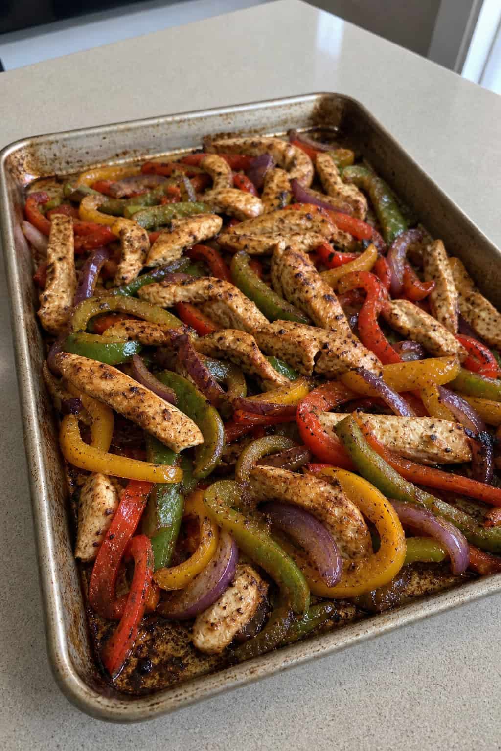 A sheet pan filled with cooked chicken breast strips, colorful bell peppers, and onions, garnished with cilantro and lime wedges.