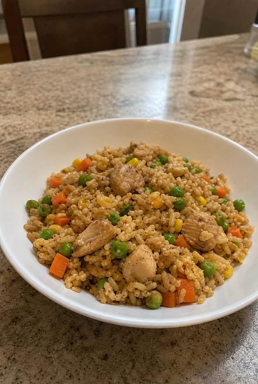 A wide bowl of golden chicken fried rice flecked with peas, carrots, corn, and green onion, with visible pieces of shredded chicken and scrambled egg throughout, photographed in a home kitchen setting.