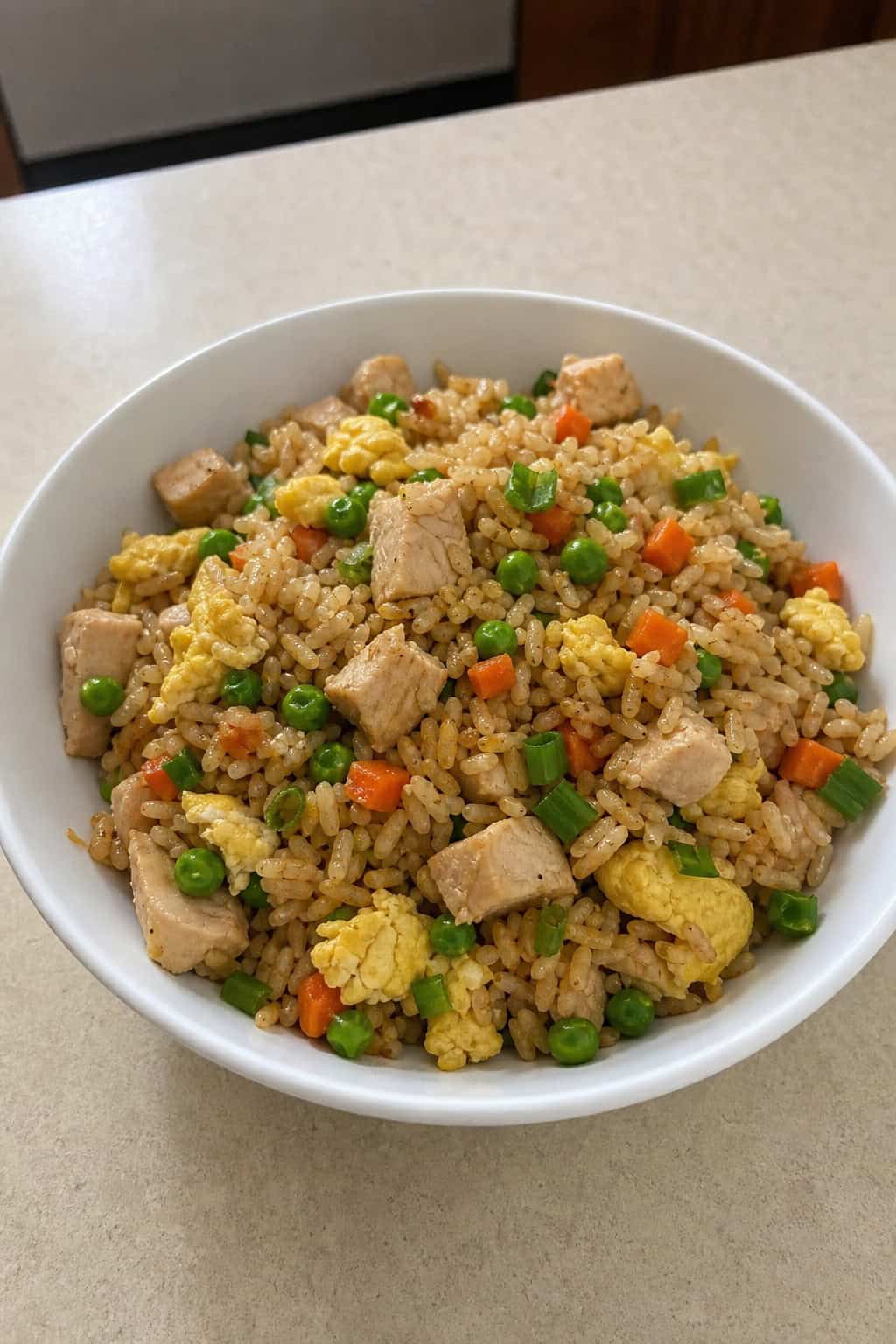 A ceramic bowl of chicken fried rice with visible peas, diced carrots, fluffy scrambled egg pieces, and sliced scallions, the rice grains separate and lightly glossed with soy sauce.