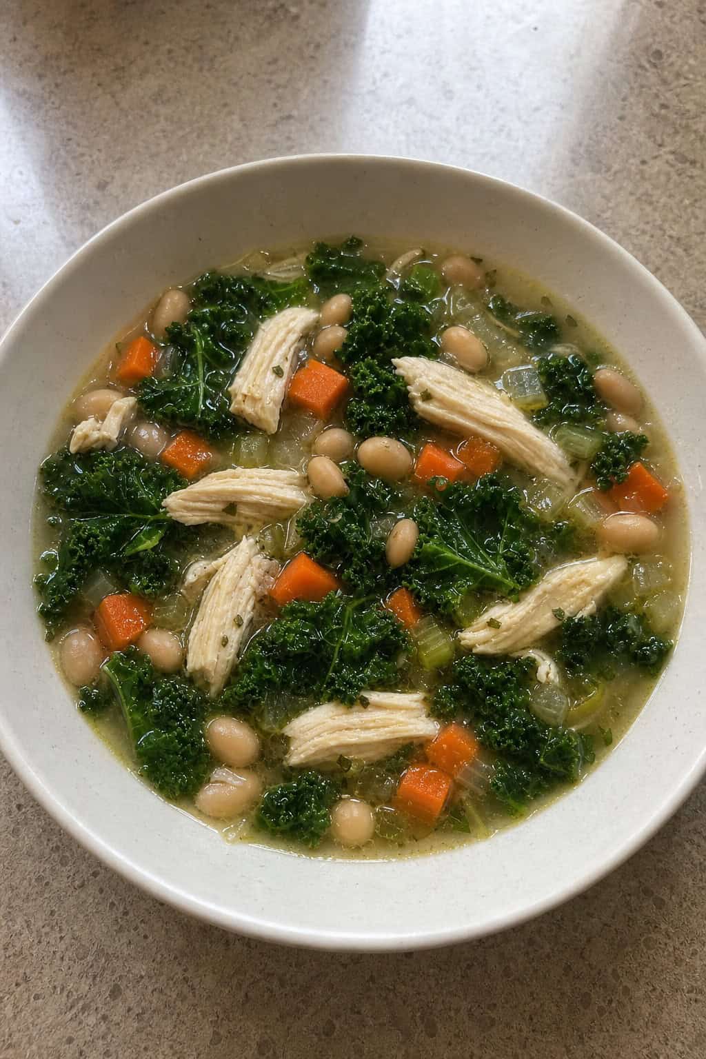 A bowl of chicken, kale, and white bean soup on a wooden table with fresh ingredients around it.