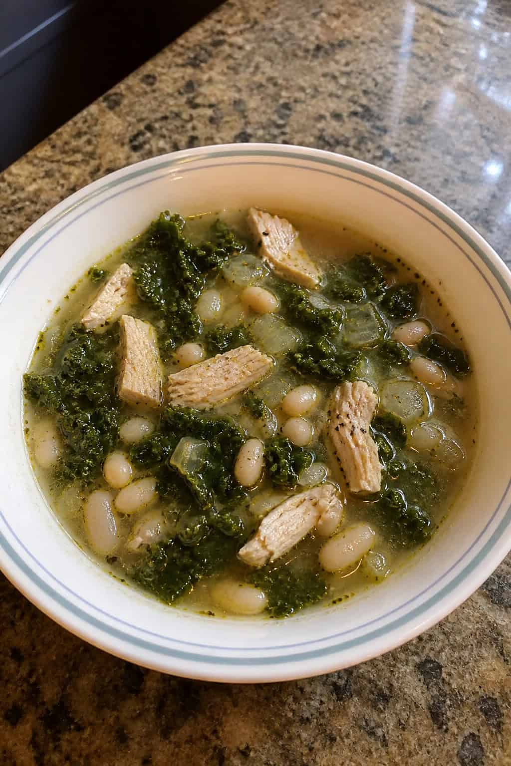 A bowl of chicken, kale, and white bean soup on a wooden table with fresh ingredients around it.