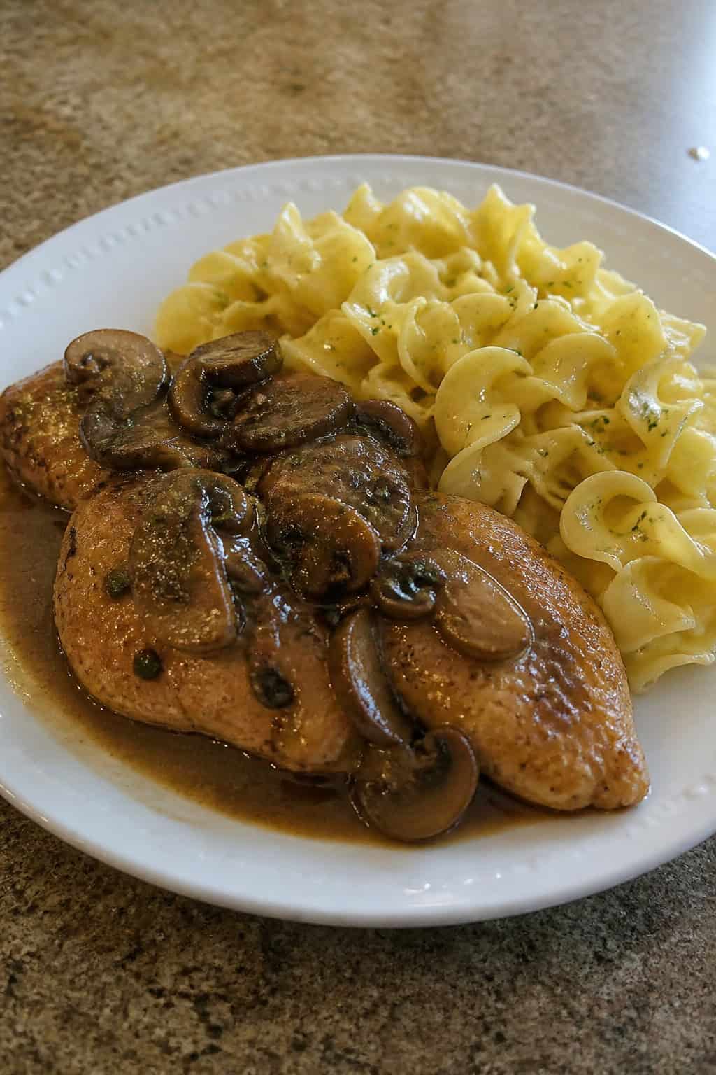 A plate of Chicken Marsala with mushroom sauce served alongside buttered noodles on a wooden table.
