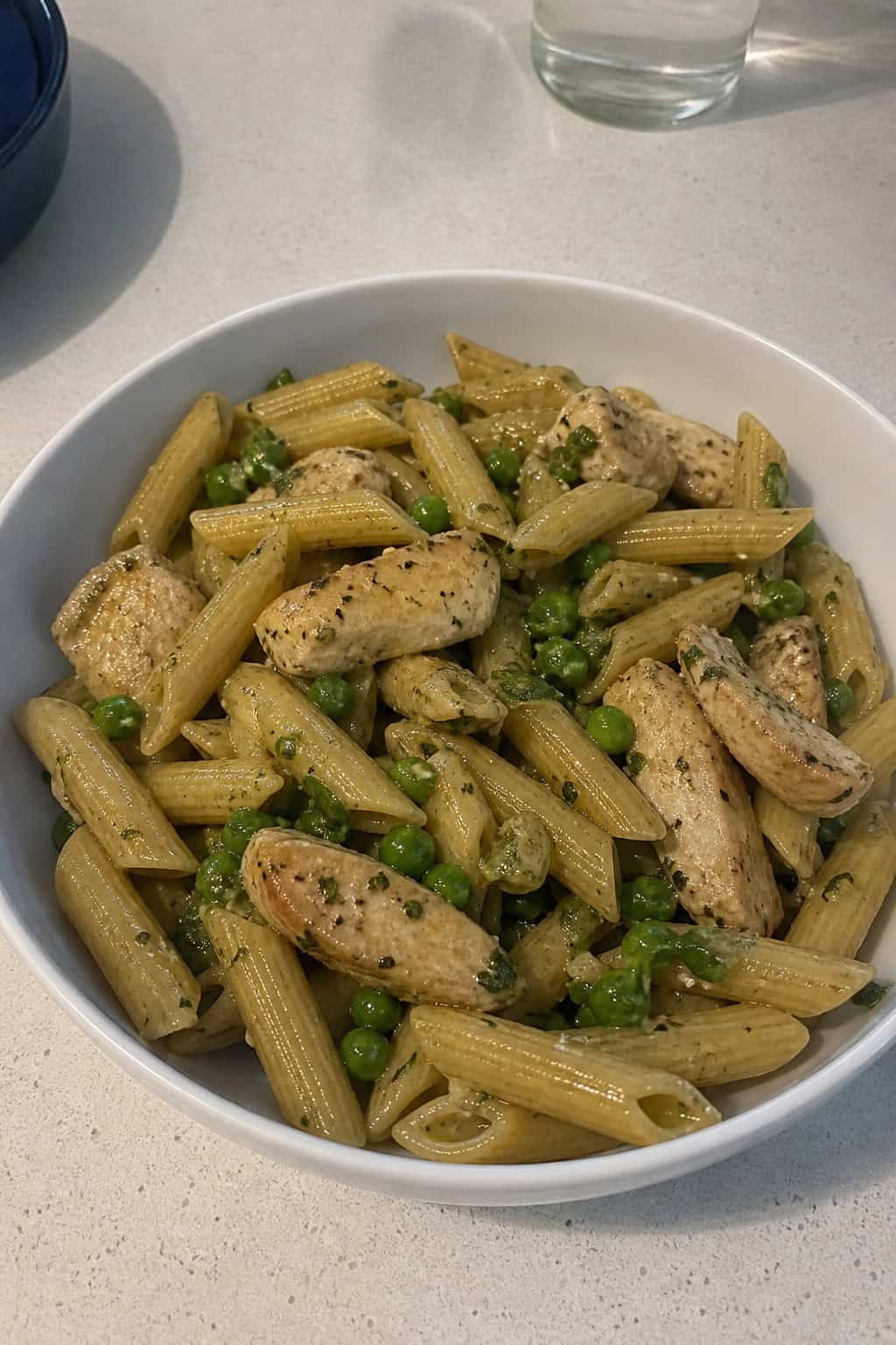 A plate of pasta with grilled chicken, peas, and mint leaves on a kitchen table with fresh ingredients in the background.