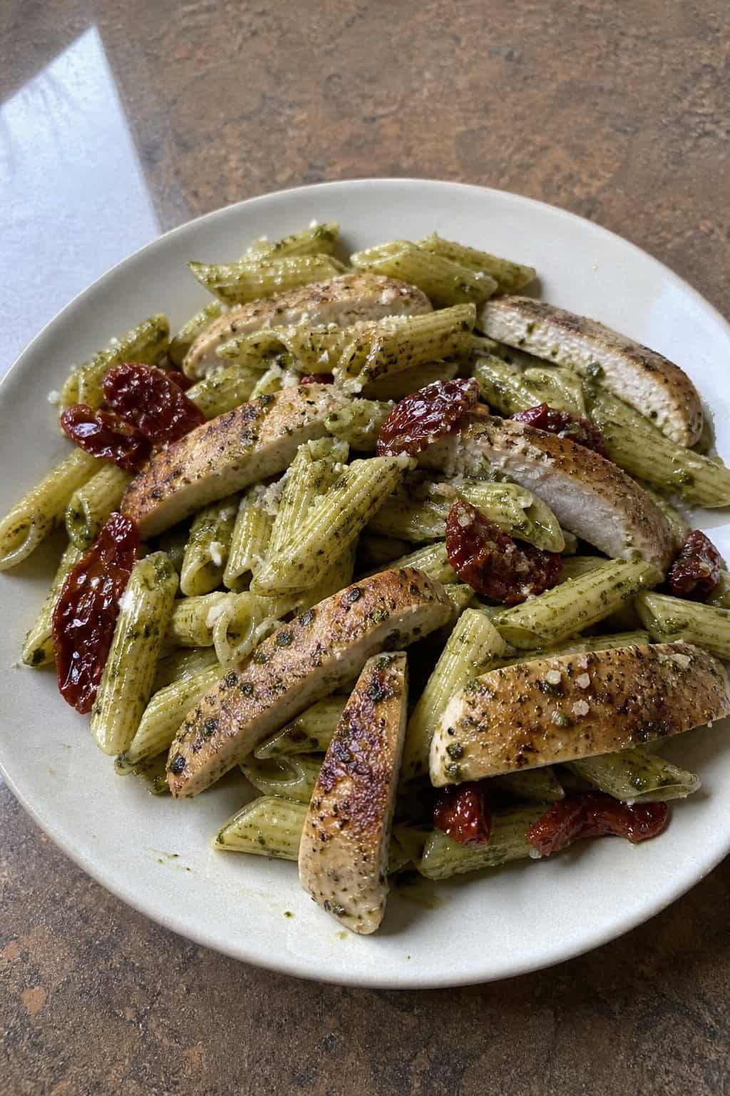 A white plate of rotini pasta coated in bright green pesto with shredded chicken and dark red sun-dried tomatoes scattered throughout, on a wooden table.