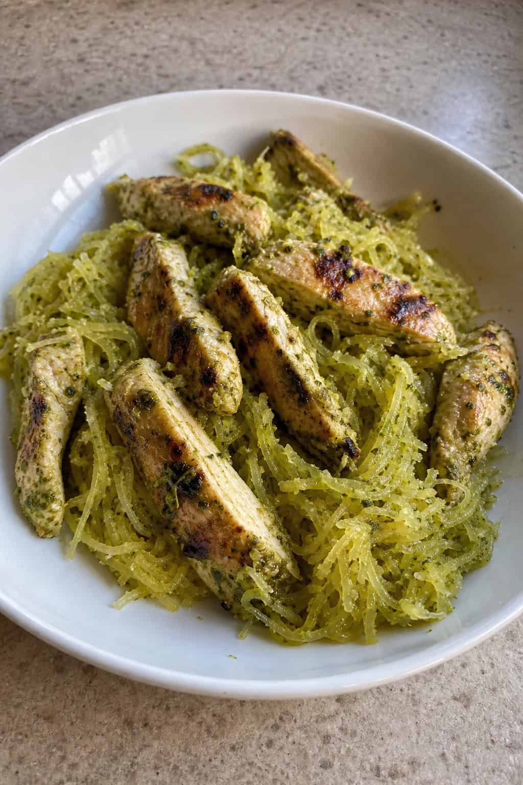 A bowl of grilled chicken mixed with spaghetti squash and green pesto sauce, garnished with basil and Parmesan cheese.
