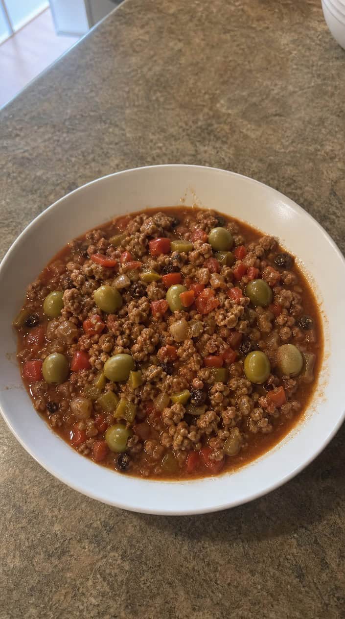 A bowl of chicken picadillo with ground chicken, green olives, raisins, and vegetables on a wooden table with fresh ingredients nearby.