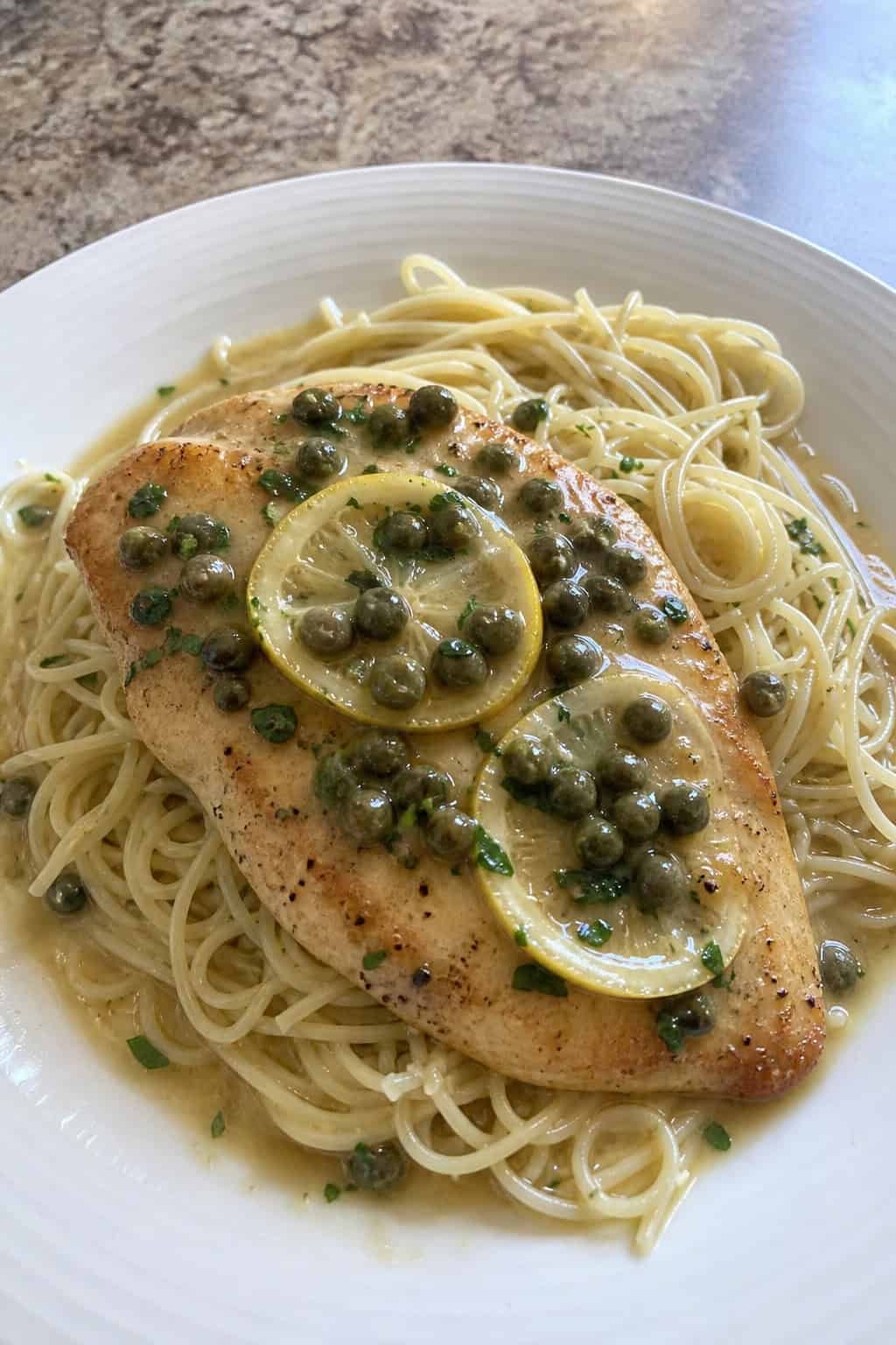 A plate of chicken piccata with angel hair pasta garnished with lemon sauce and parsley on a wooden table.