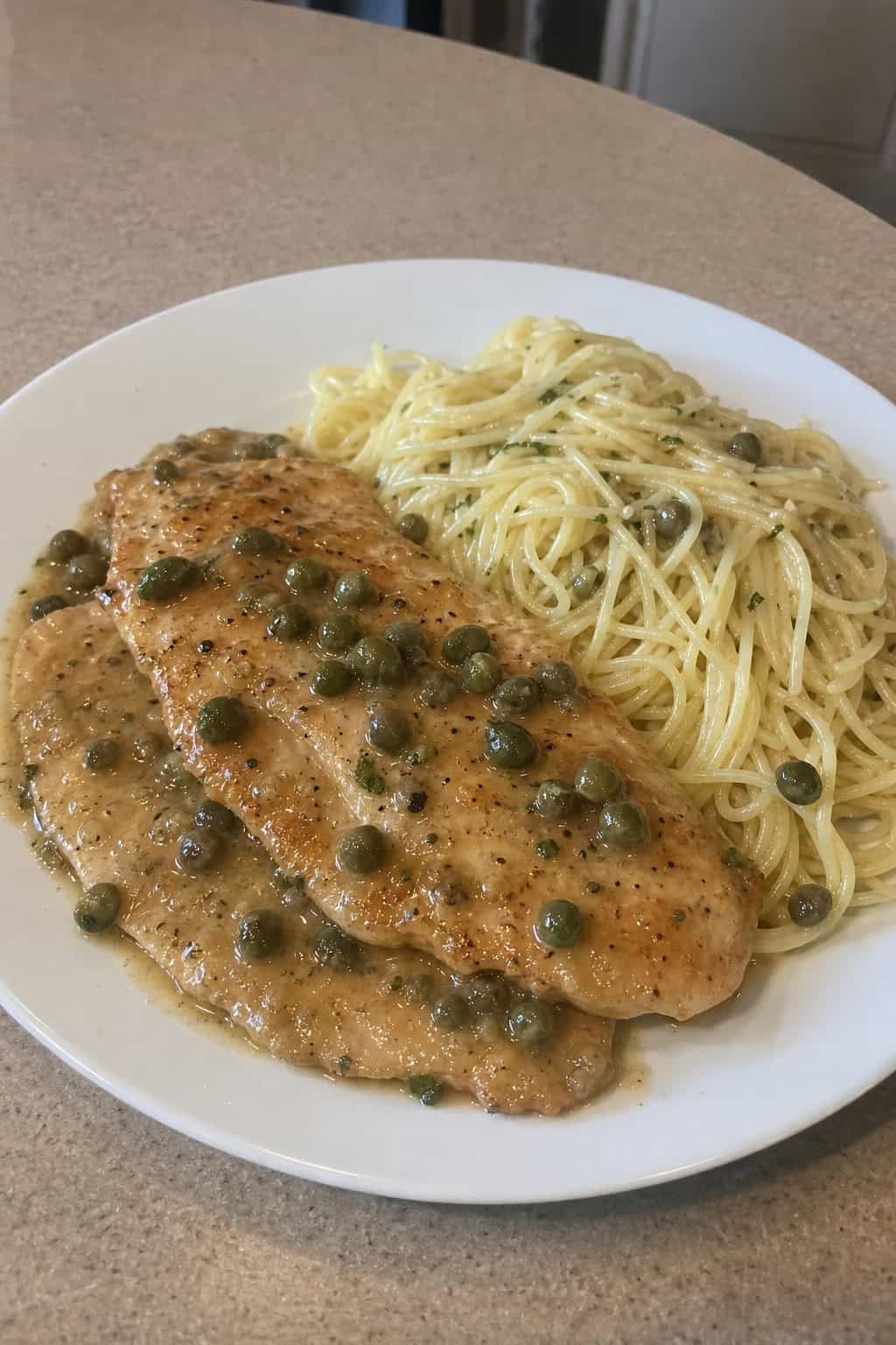 A plate of Chicken Piccata served with angel hair pasta, garnished with lemon slices and fresh parsley on a wooden table.