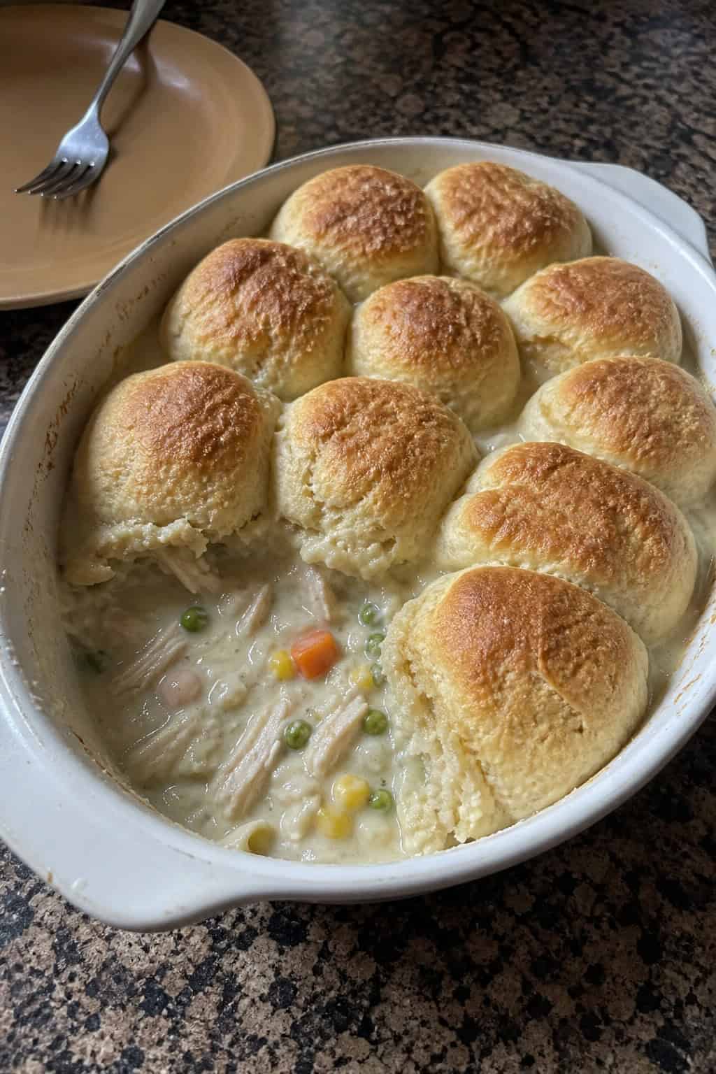 A freshly baked chicken pot pie casserole with tall, golden biscuit rounds on top, steam rising, on a wooden table with a serving spoon and fresh herb sprigs nearby.