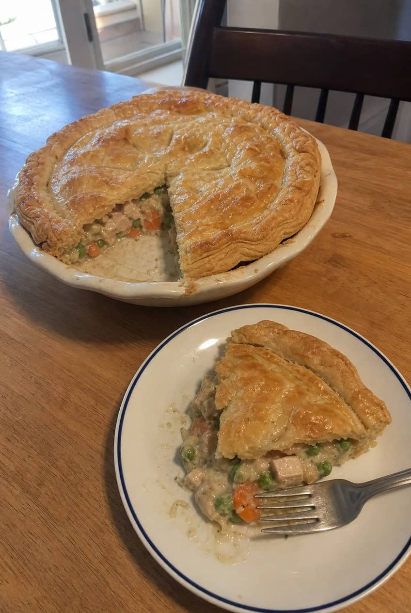 A deep golden-brown chicken pot pie with a flaky puff pastry crust, partially sliced to reveal a creamy vegetable and chicken filling inside, set on a wooden table with a serving plate and fresh vegetables in the background.