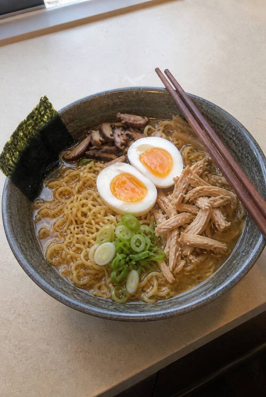 A deep bowl of homemade-style chicken ramen with springy ramen noodles in a dark soy broth, topped with a halved soft-boiled egg with a jammy yolk, scattered green onions, wilted spinach, and shredded rotisserie chicken, with chopsticks resting on the rim.