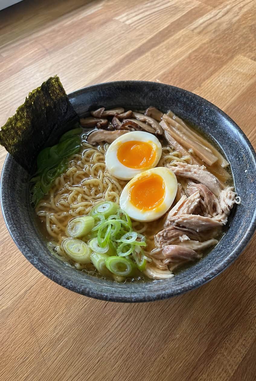 A deep bowl of homemade-style chicken ramen with springy ramen noodles in a dark soy broth, topped with a halved soft-boiled egg with a jammy yolk, scattered green onions, wilted spinach, and shredded rotisserie chicken, with chopsticks resting on the rim.