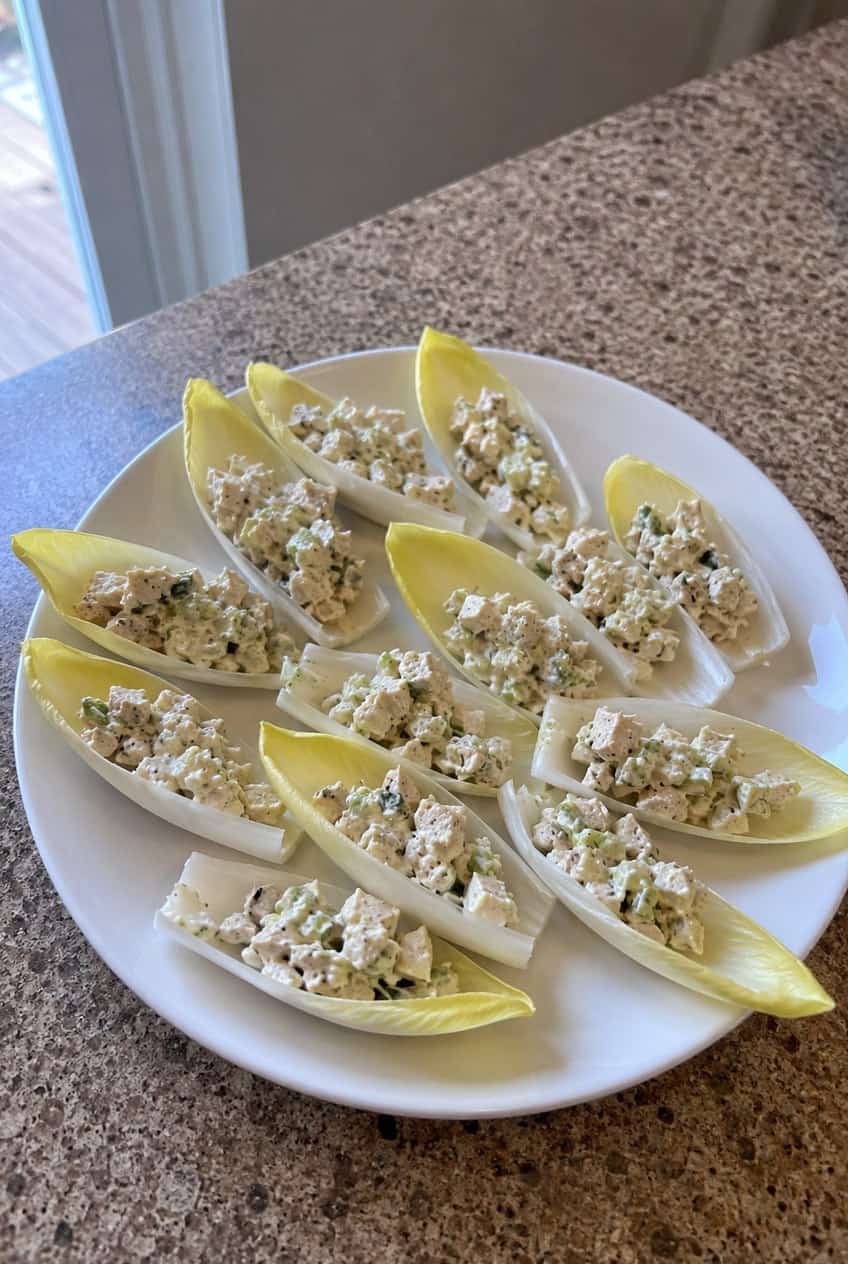 A wooden board with pale yellow Belgian endive leaves filled with a creamy, chunky chicken salad, surrounded by scattered fresh herb sprigs.
