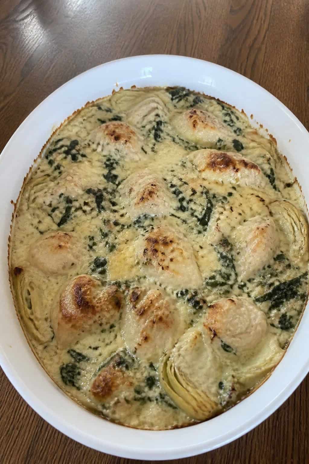 A baked chicken casserole with chopped spinach and artichoke hearts in a cream cheese base, topped with a golden panko crust in a ceramic dish on a wooden table.