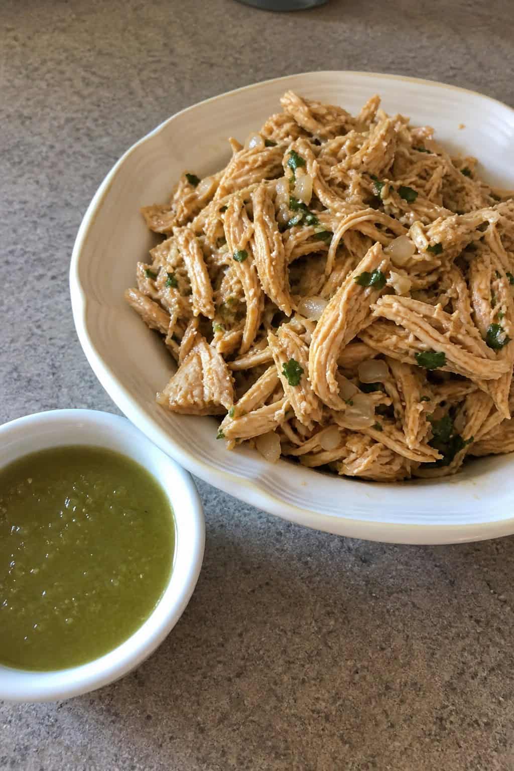 A bowl of shredded chicken taco filling next to a dish of green salsa verde with fresh lime wedges and herbs on a wooden surface.
