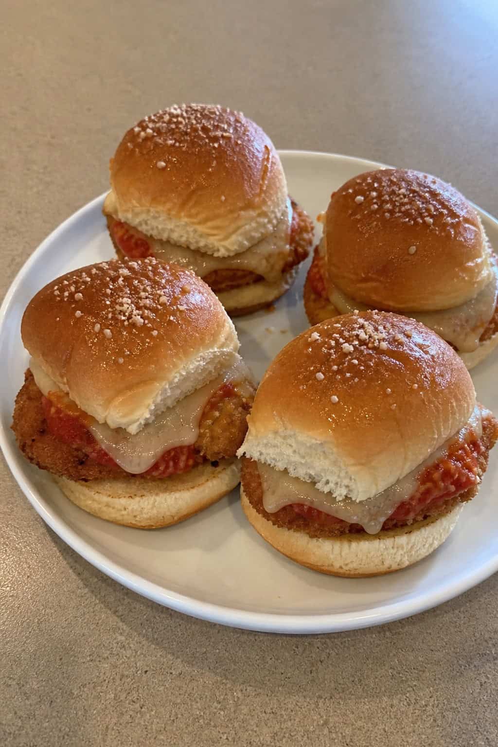 A plate of chicken tender parmesan sliders with melted cheese and marinara sauce on slider buns, garnished with lettuce and basil on a wooden table.