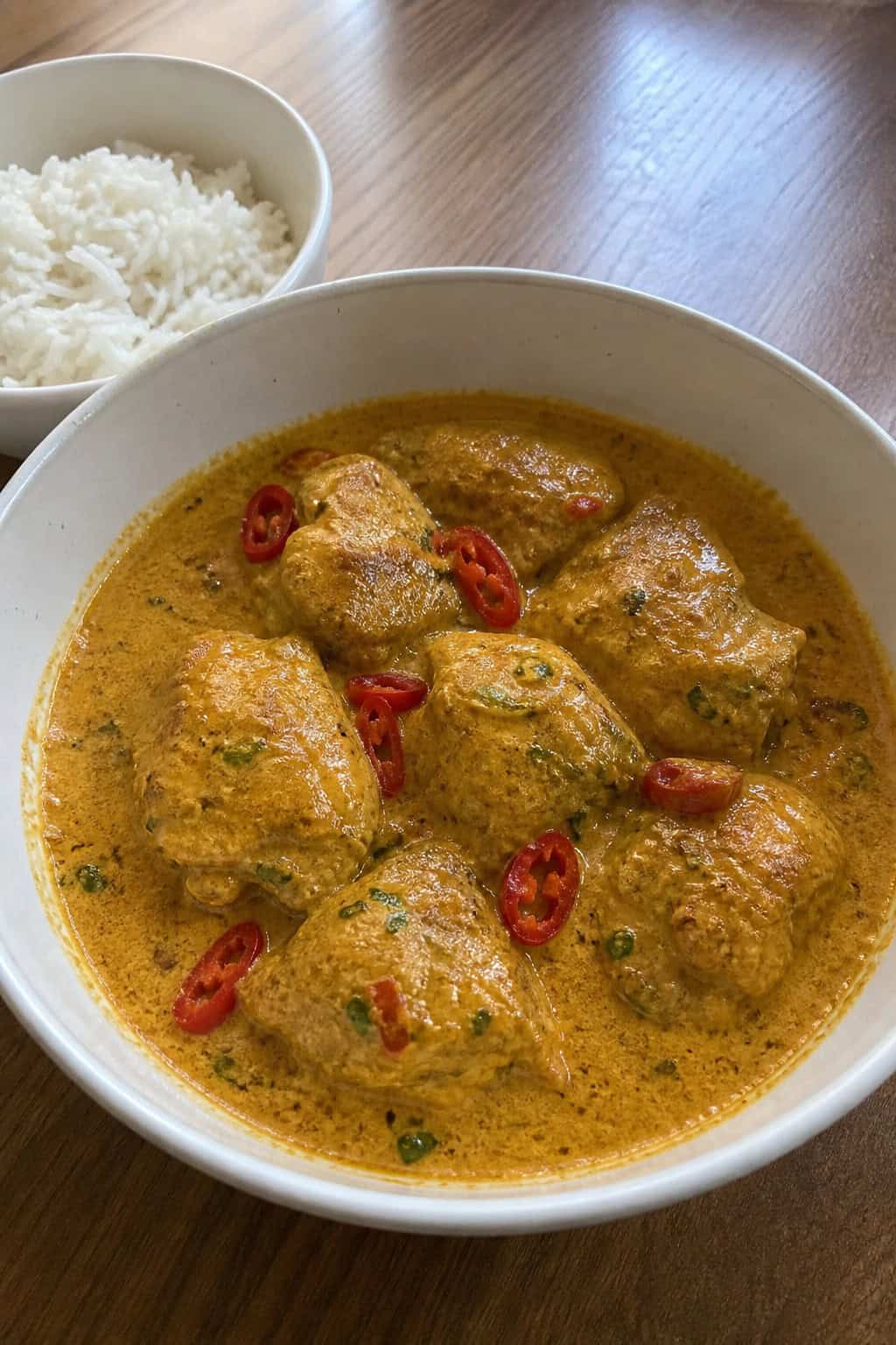 A wide bowl of bite-sized chicken thighs in a deep golden coconut curry sauce, garnished with fresh cilantro and thin red chili slices, with cooking ingredients visible on the wooden table behind it.