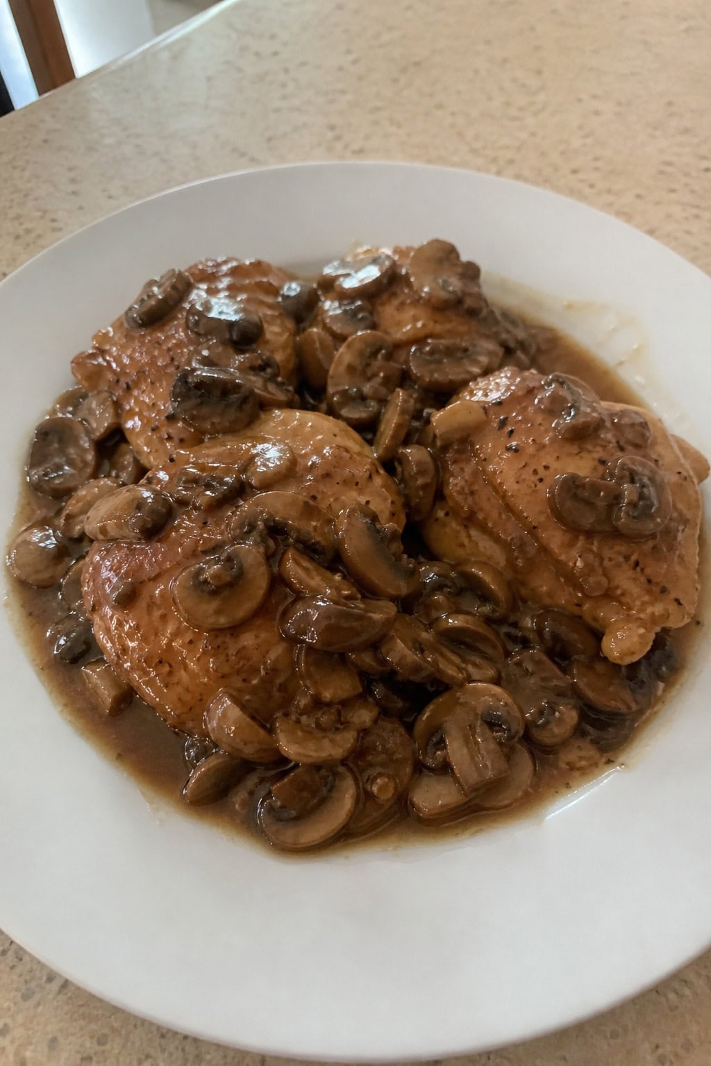 Plate of golden brown chicken thighs covered in mushroom Marsala sauce, garnished with parsley, on a wooden table with a glass of red wine nearby.