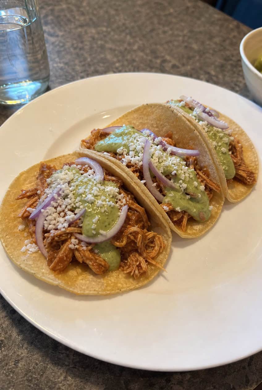 A ceramic plate holding three corn tortilla tacos filled with smoky chicken tinga, topped with a pale green avocado crema, fresh cilantro, and thinly sliced red onion, with lime wedges arranged alongside on a wooden table.