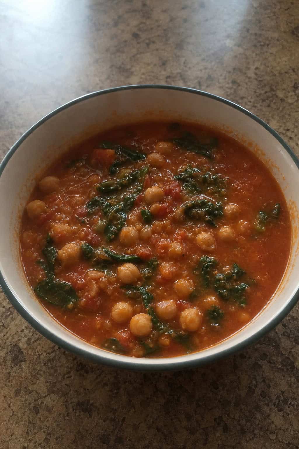 A bowl of chickpea, tomato, and spinach soup on a wooden table with fresh ingredients around it.