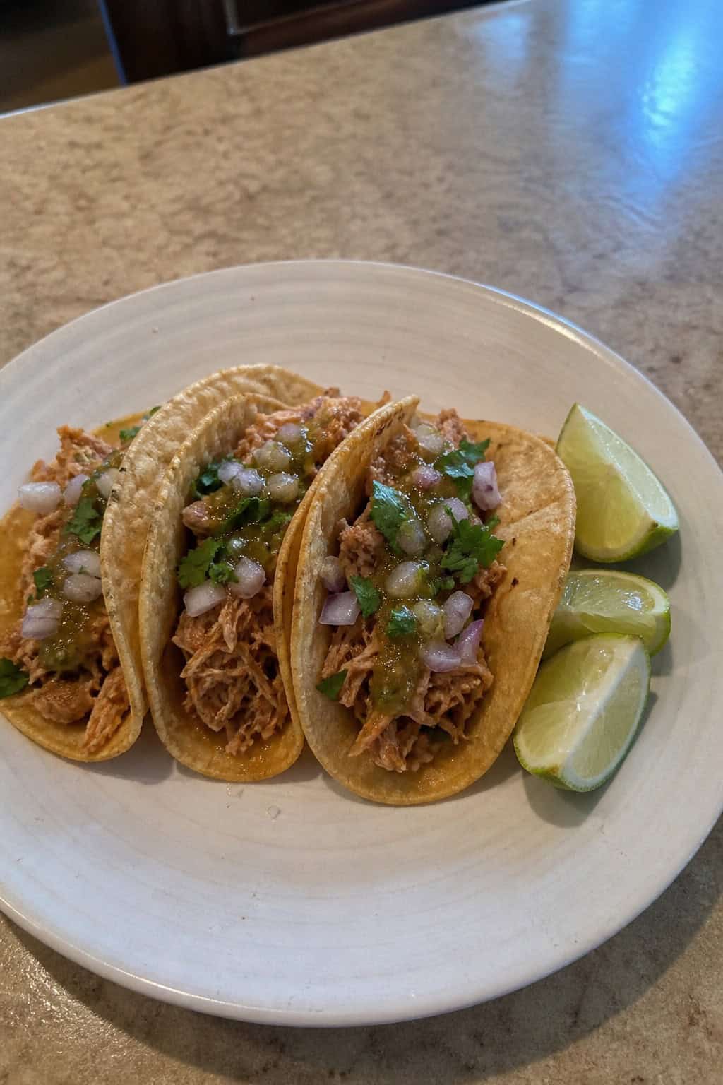Three chili lime chicken tacos on a plate with lime wedges and fresh cilantro on a wooden table.