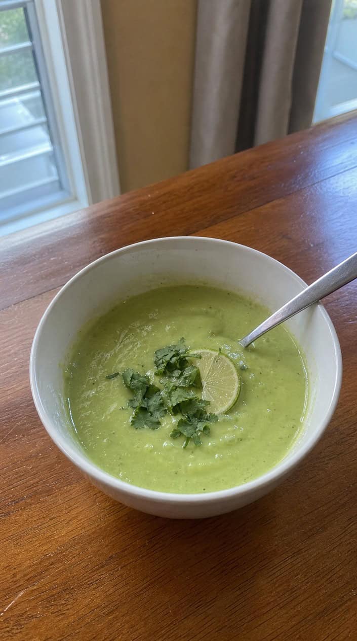 A bowl of chilled avocado-cilantro soup garnished with cilantro and lime wedges on a wooden table with fresh avocados, cilantro, and limes around it.