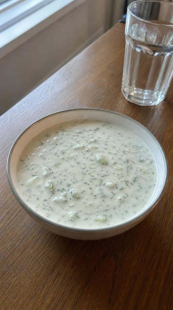A bowl of chilled cucumber and dill soup with fresh ingredients on a wooden table.
