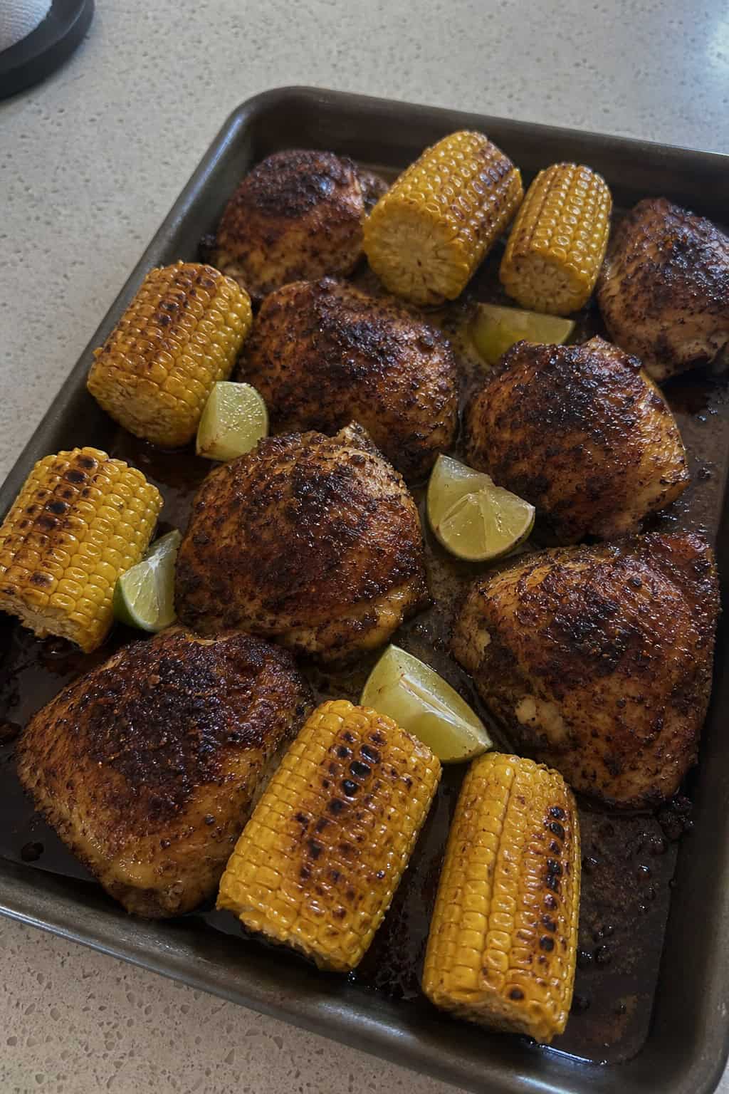A foil-lined sheet pan with chipotle-lime chicken thighs roasted alongside charred corn kernels, sliced red bell pepper, and red onion, with lime wedges and cilantro scattered on top.