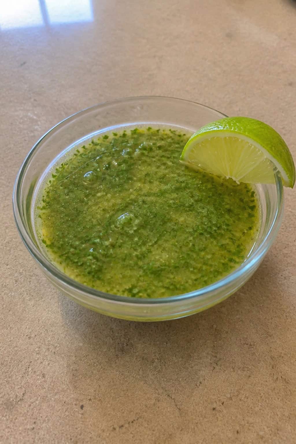 A bowl of green cilantro-lime vinaigrette surrounded by fresh cilantro, limes, and garlic on a wooden table.